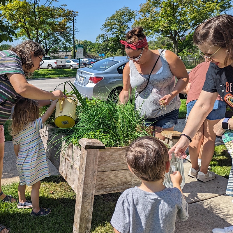 Adults and children tend a raised garden bed outside near a sidewalk. One adult helps a child use a watering can.