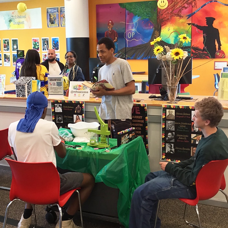 Two teens sit at a table in the library and a third stands, holding a book