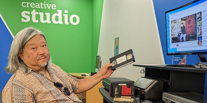 Person holds a VHS tape at a computer station with the words "Creative Studio" on the wall