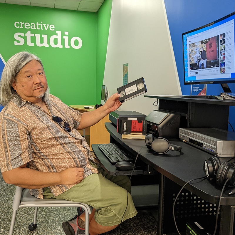 Man holds up VHS tape at computer station, with words Creative Studio on wall