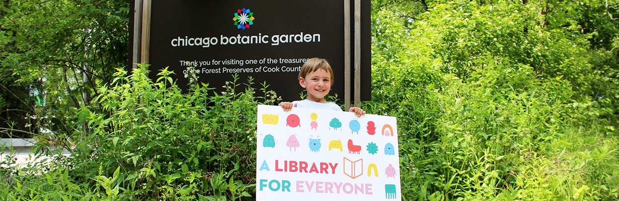 Young boy holding a library card in front of the Chicago Botanic Garden