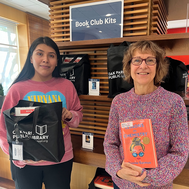 Two library staff hold a bag and a book next to a shelf that says Book Club KIts