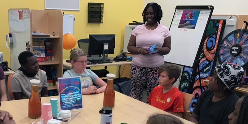 Group of middle schoolers sit around a table and talk with each other and an adult who's standing up