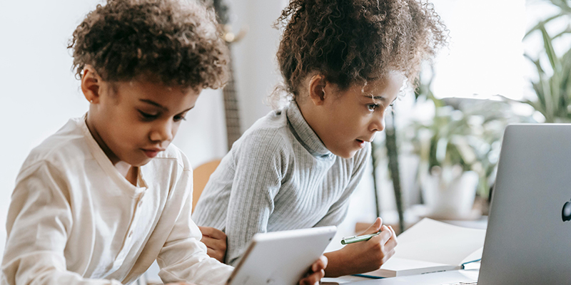 Two kids sitting next to each other, one on a tablet and the other using a laptop